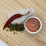 Tin of mild chilli seaweed salt on a wooden board with sea salt, dried seaweed and a fresh red chilli 